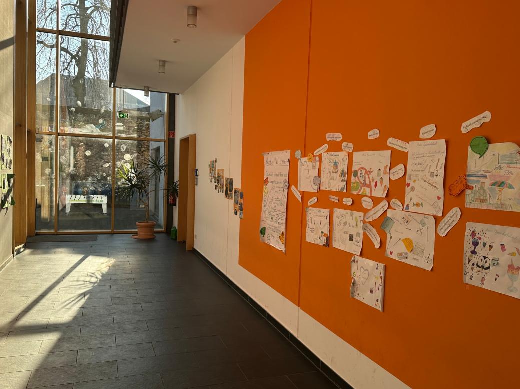 Man schaut in den Flur im Erdgeschoss der Grundschule am Dom. An der rechten Flurseite sind Erinnerungsplakate zu sehen. Am Flurende ist eine Glaswand zum Schulhof.