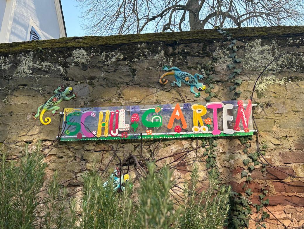 Man sieht das Wandbild 'Schulgarten', das an der Mauer hinter dem Schulgarten angebracht ist.