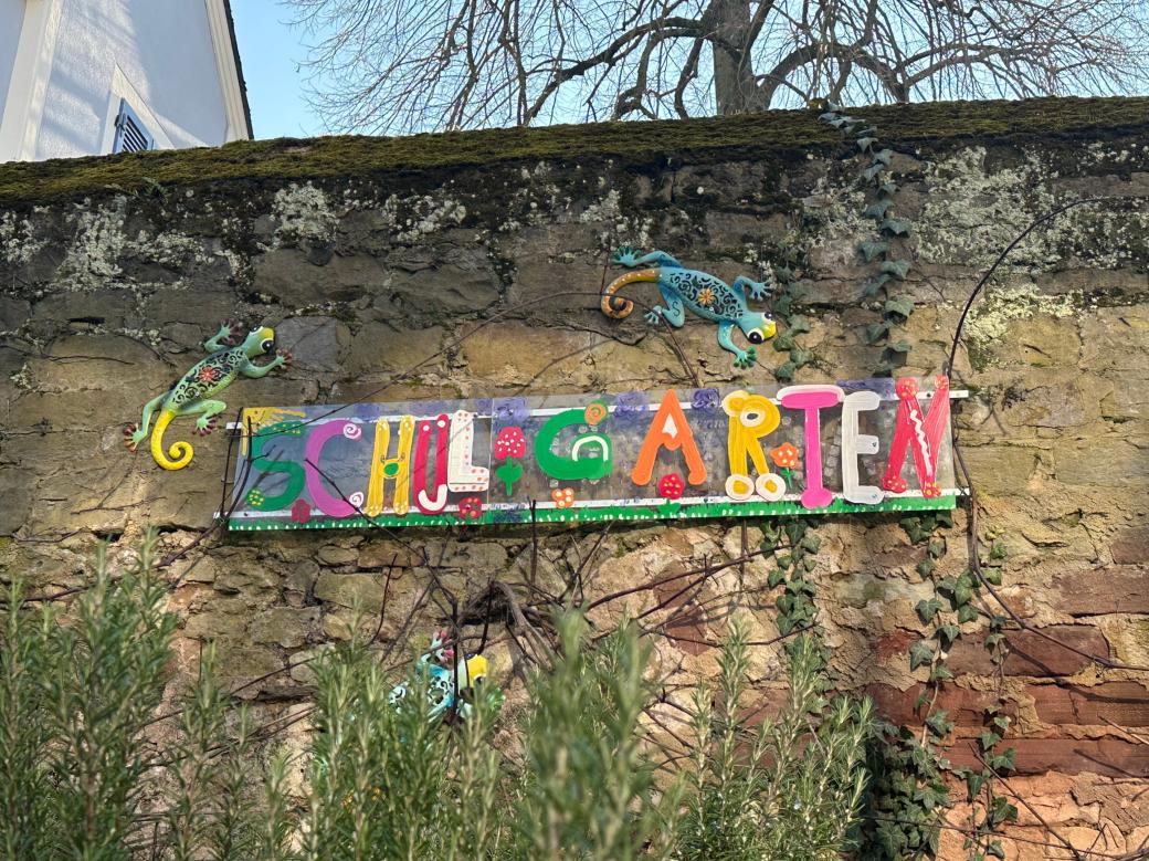 Man sieht das Wandbild 'Schulgarten', das an der Mauer hinter dem Schulgarten angebracht ist.