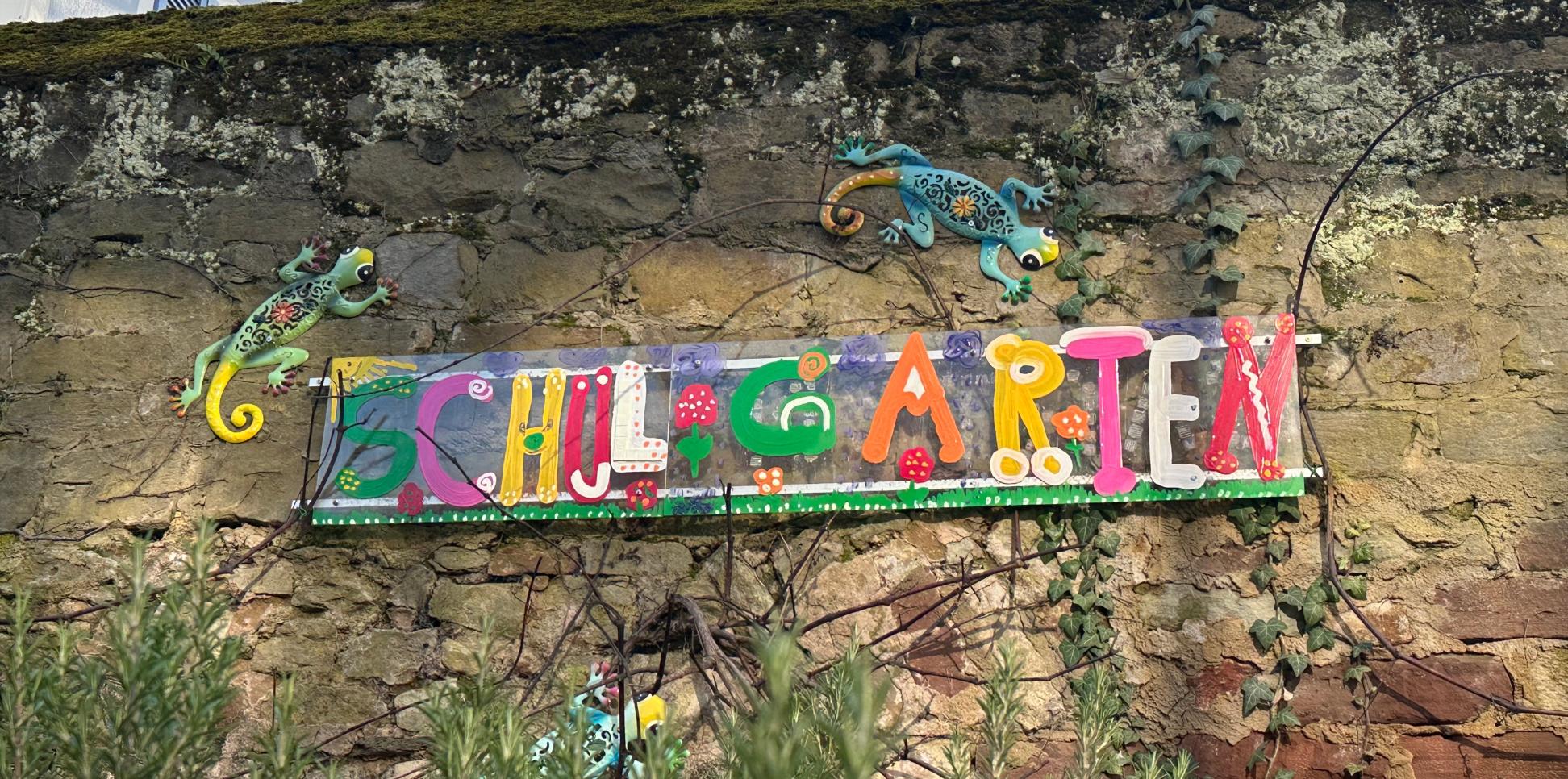Man sieht das Wandbild 'Schulgarten', das an der Mauer hinter dem Schulgarten angebracht ist.
