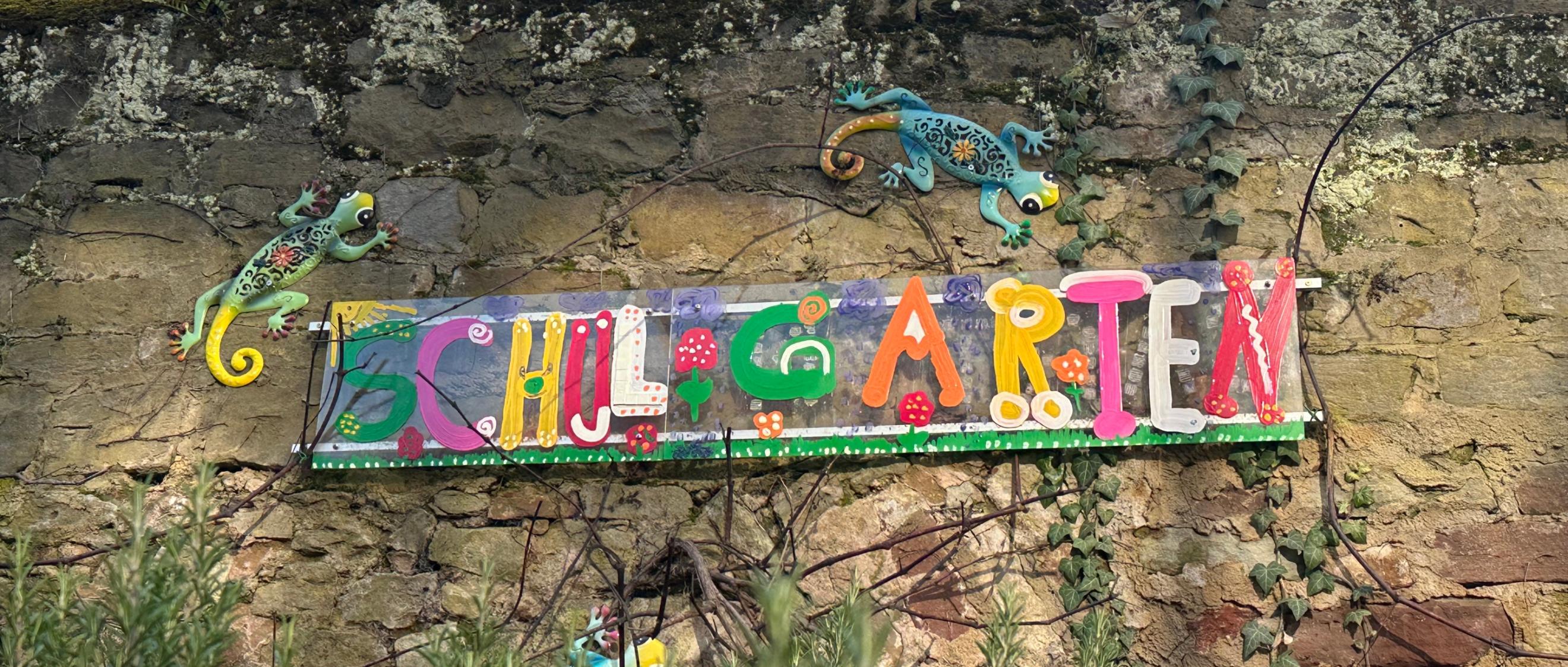Man sieht das Wandbild 'Schulgarten', das an der Mauer hinter dem Schulgarten angebracht ist.