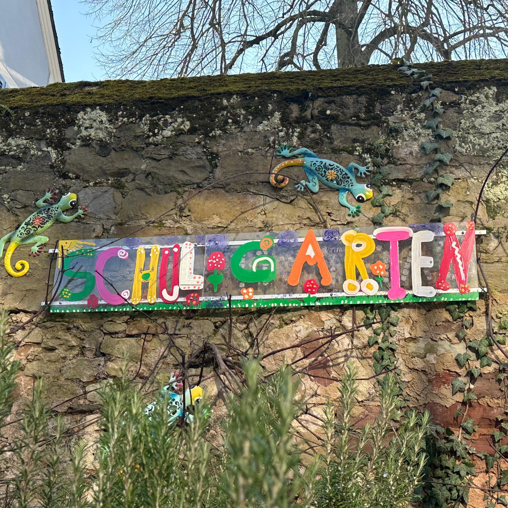 Man sieht das Wandbild 'Schulgarten', das an der Mauer hinter dem Schulgarten angebracht ist.