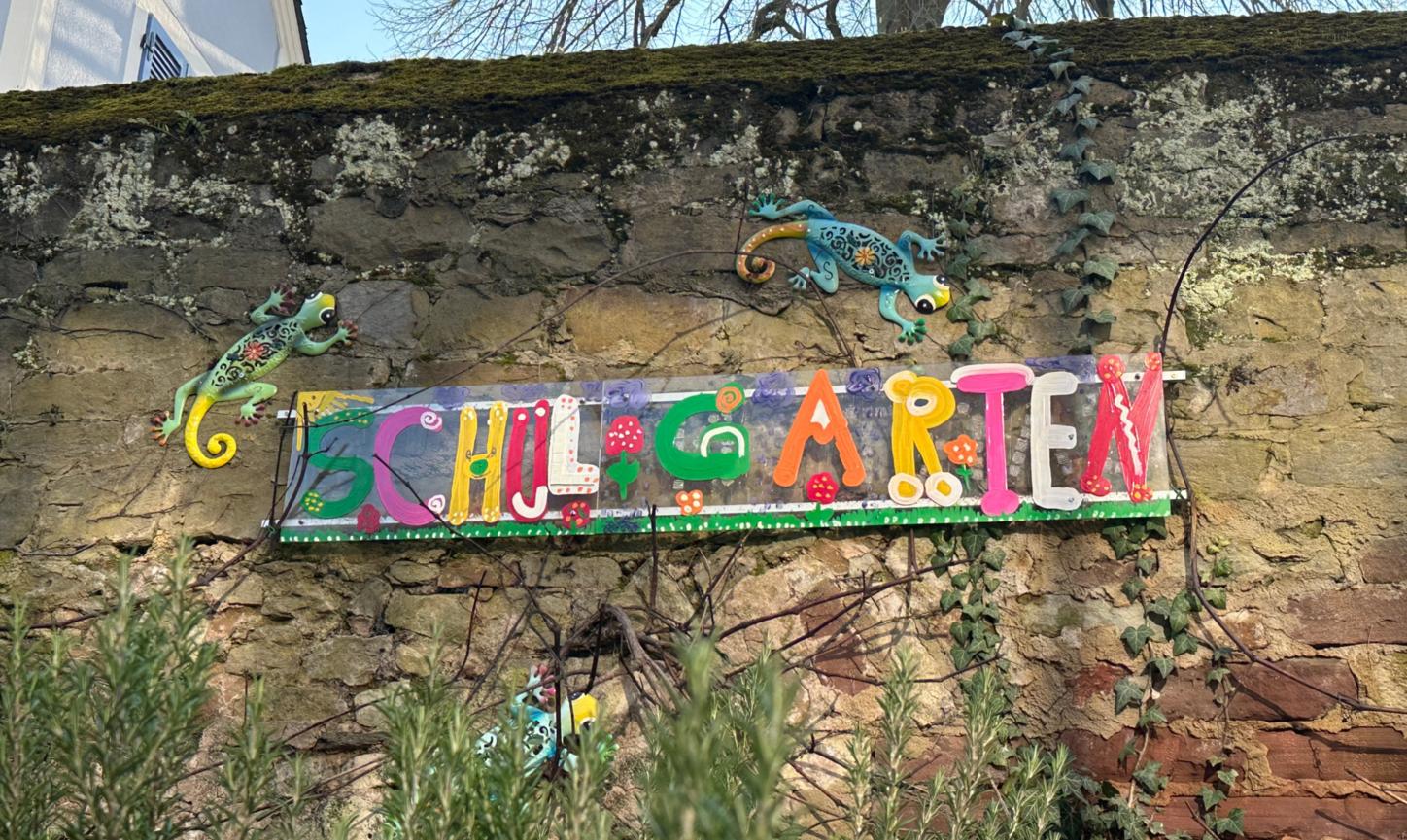 Man sieht das Wandbild 'Schulgarten', das an der Mauer hinter dem Schulgarten angebracht ist.